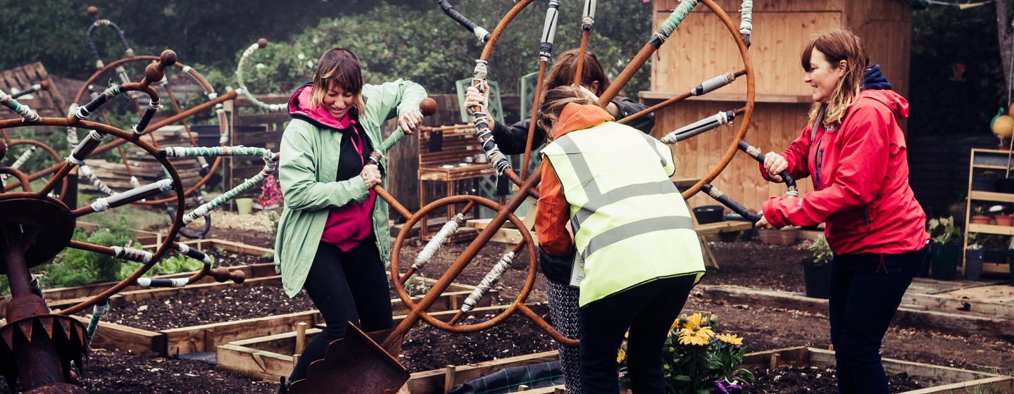 'Our Common Tools' by Katie Surridge b-side festival 2021. The photo depicts 4 people in an allotment carrying a very large tool like a shovel 