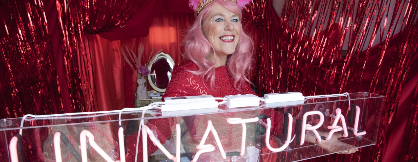 An image of a woman standing in front of a glitter fringe curtain decorated beauty salon, with a neon sign saying 'Unnatural"
