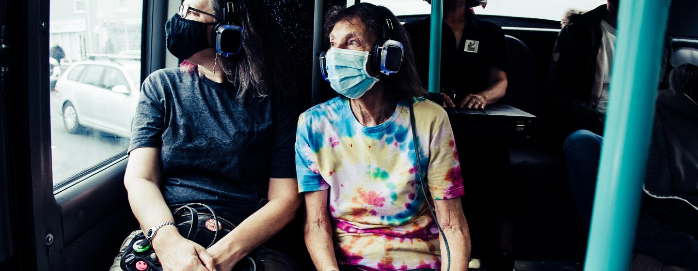 An image of two people sitting on a bus looking out of the window, with a headset on