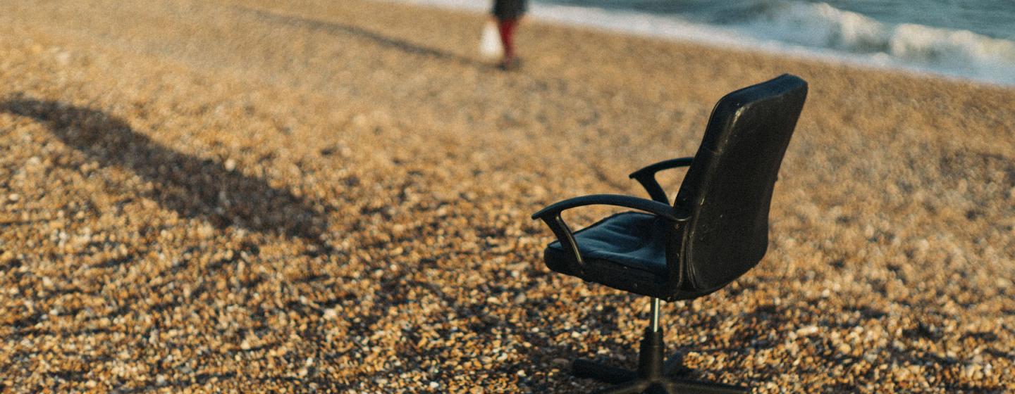 An image of a chair on a pebble beach with the sea in the background and a person walking past