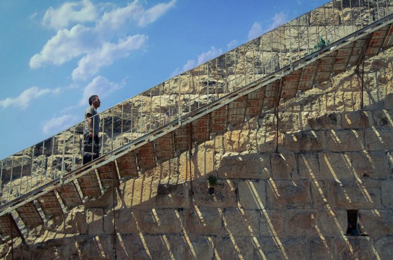 An image of a man walking up a set of stairs with a wall and the blue sky behind him 