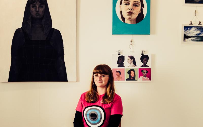 An image of a girl with a pink top on, sitting down in front of an exhibition of paintings.