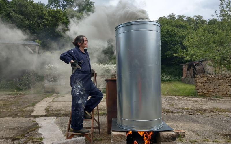 A woman in a boiler suit stands next to a metal chimney with smoke